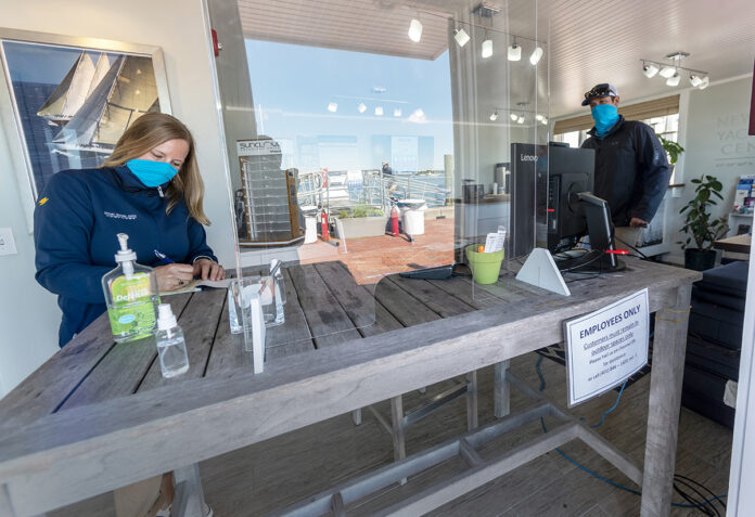 NEW ARRANGEMENTS: Newport Yachting Center Office Manager Leanne Arsenault, left, is no longer using her regular office desk. Now she greets visitors from a makeshift desk at the door with a plexiglass shield. At right is Carter Fisher, Newport Yachting Center dock master. / PBN PHOTO/MICHAEL SALERNO
