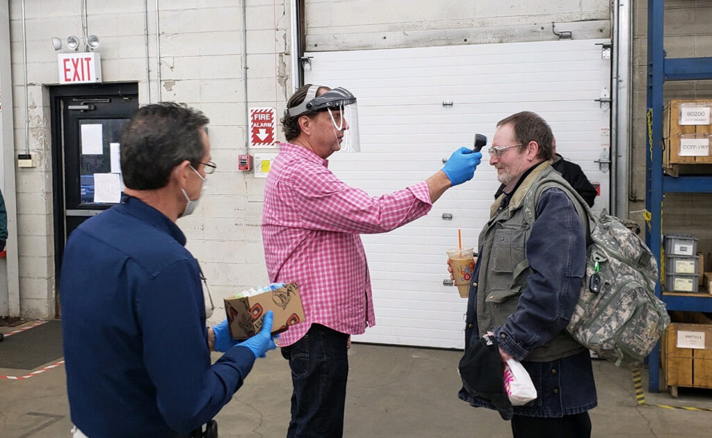 DAILY CHECK UP: David Bragonier, VIBCO machinist and value stream manager, has his temperature measured by CEO and President Karl Wadensten as Bragonier enters the factory. At left, production manager John Goodwin holds a box of hand sanitizer. When this photo was taken, facemasks were not yet required. / COURTESY VIBCO INC.