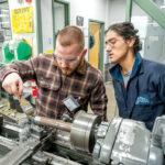 SPIN CYCLE: New England Institute of Technology Advanced Manufacturing Coordinator Todd Sposato, right, works with student Zach Carmody, 24, of Charlestown on the lathe at the Shipbuilding/Marine and Advanced Manufacturing Institute workroom at New England Tech’s Warwick campus.  PBN PHOTO/DAVE HANSEN