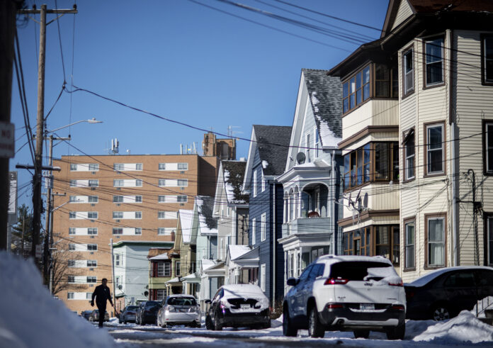 A MAN WALKS to clean snow off a car along a row of homes in Central Falls, the community hit hardest by the pandemic in the state. / AP FILE PHOTO/DAVID GOLDMAN A MAN WALKS to clean snow off a car along a row of homes in Central Falls, the community hit hardest by the pandemic in the state. / AP FILE PHOTO/DAVID GOLDMAN
