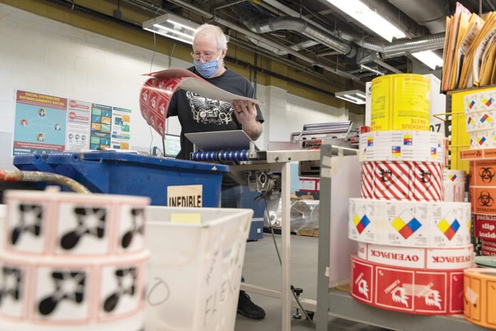 SIGNS OF THE TIMES: National Marker Co., creator of safety identification signs and products, increased its sales by more than 25% in 2020, as its products were in high demand during the COVID-19 pandemic. At right, manufacturing technician Tim Egan looks over some signs. / PBN PHOTO/RUPERT WHITELEY 