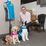LAB MODEL: Steven Triedman, co-owner of Corky’s Reflective Wear LLC, which makes highly visible and water-repellent collars and jackets for dogs, with Georgie, a yellow Lab puppy modeling the K-9 reflective wear, flanked by two stuffed models. / PBN PHOTO/MICHAEL SALERNO