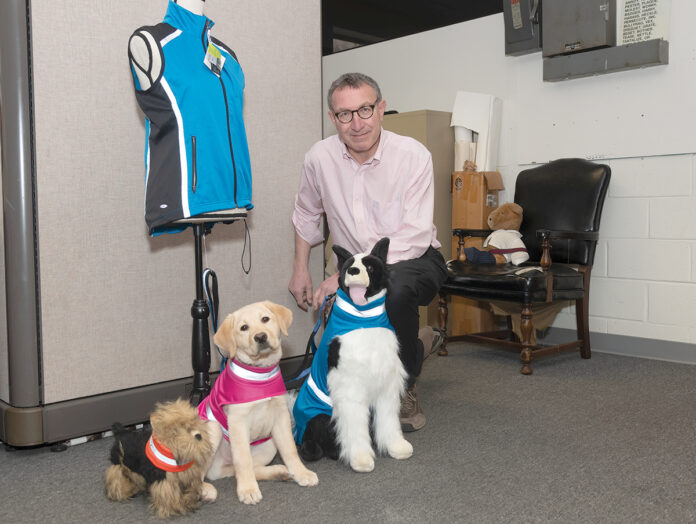 LAB MODEL: Steven Triedman, co-owner of Corky’s Reflective Wear LLC, which makes highly visible and water-repellent collars and jackets for dogs, with Georgie, a yellow Lab puppy modeling the K-9 reflective wear, flanked by two stuffed models. / PBN PHOTO/MICHAEL SALERNO LAB MODEL: Steven Triedman, co-owner of Corky’s Reflective Wear LLC, which makes highly visible and water-repellent collars and jackets for dogs, with Georgie, a yellow Lab puppy modeling the K-9 reflective wear, flanked by two stuffed models. / PBN PHOTO/MICHAEL SALERNO