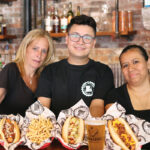 THE WORKS: From left, Harry’s Bar & Burger staff members Lynn Perrino, Kelvin Agustin and Sonia Real display Gastros Craft Meats hot dogs at the restaurant’s Atwells Avenue location in Providence. The comfort food is on the Harry’s menu at all locations. / COURTESY CHRISTINA MONAHAN