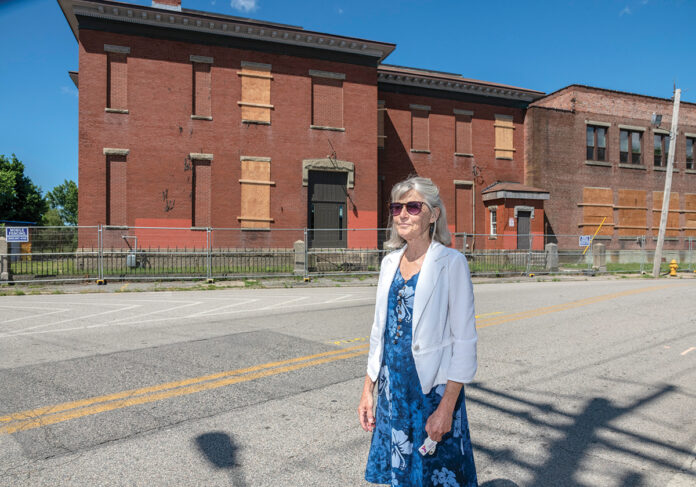 REBIRTH? Jan Brodie, executive director of the Pawtucket Foundation, stands outside an abandoned industrial building at Conant and Pine streets in Pawtucket that city officials hope will be part of the redevelopment envisioned around a commuter rail and bus station scheduled to be finished in 2022. / PBN PHOTO/MICHAEL SALERNO