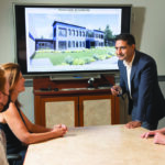 SNEAK PEEK: Windmoeller & Hoelscher Corp. Senior Vice President Javeed Buch, standing, meets with staffers at the firm’s North American headquarters in Lincoln. On the screen is an artist’s rendering of the company’s future offices that are soon to be under construction. / PBN PHOTO/DAVE HANSEN