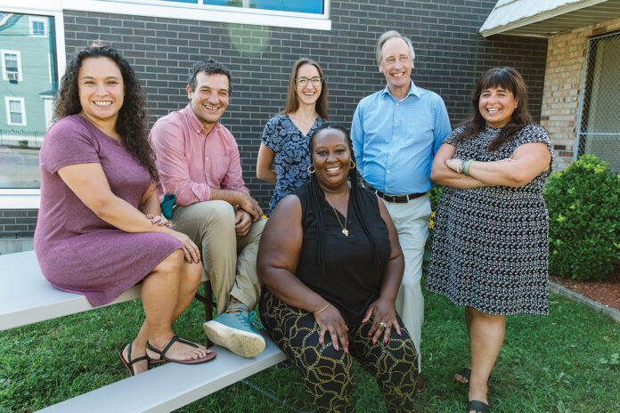 COLLABORATIVE CREW: Children’s Friend and Service employees, from left, Lucretia Lopez, enrollment supervisor; Joshua Wizer-Vecchi, innovation manager; Shelby Mack, standing, strategy manager; Owen Heleen, director of program development and innovation; Stacy Couto, chief of philanthropy; and Rachel Cooper, sitting middle, manager of family preservation, gather outside the organization’s Providence office. / PBN PHOTO/RUPERT WHITELEY