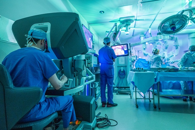 DR. JORDAN F. HEBERT, seated left, manipulates the robotic arms of the da Vinci Xi from a console in the operating room to implant the LINX device at South County Hospital. / COURTESY SOUTH COUNTY HEALTH