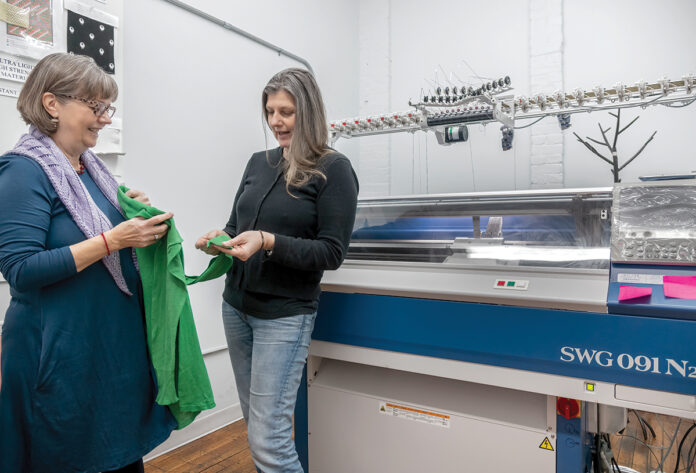 PRACTICAL ­ATTIRE: Propel LLC owner Clare King, left, and Birgit Leitner, product design and development lead, examine an aircraft carrier flight deck jersey Propel manufactures for the U.S. Navy. The company’s whole-garment knitting machine is to the right. / PBN PHOTO/MICHAEL SALERNO
