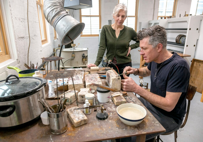 MALLEABLE METALLERS: Sandi Bonazoli and Jim Dowd are the co-owners of Beehive Handmade, a long-standing metal­work business that recently opened a store and studio space in Warren. Dowd crafts a copper rack for measuring spoons while Bonazoli looks on. / PBN PHOTO/MICHAEL SALERNO