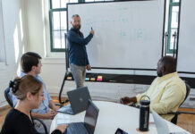 BOARD MEETING: Marstone Inc. Senior Solutions Architect Ken Linehan, standing, leads a team meeting at the Providence office. Also part of the meeting are, from left, Office Manager Caiti Moran, Director of Sales Ben Baker and digital marketing strategist Farouk Ajakaiye.  PBN PHOTO/TRACY JENKINS