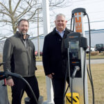 AMPED UP: Zachary Cobb, left, Hexagon Manufacturing Intelligence vice president of services, and Steven Ilmrud, vice president of operations, demonstrate one of the charging stations at the North Kingstown company that are part of the company’s ambitious plans to offset the emissions it produces with renewable energy or credits by 2030.  PBN PHOTO/ELIZABETH GRAHAM
