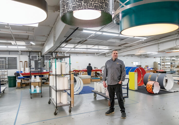 SEEING THE LIGHT: Ian Prichett, CEO and president of Lumetta Inc., stands on the floor of the Warwick lighting manufacturer with some of its designs hanging overhead. He says technologies and product designs evolve quickly in the lighting world, so he’s constantly spending time on the floor and learning from each ­department. PBN PHOTO/MICHAEL SALERNO