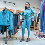 WHAT’S IN STORE: Toyin Omisore, owner of Roam Loud, an activewear and athleisure brand, stands with the company’s products on display at REI in Cranston. Omisore has also sold her products through Kohl’s Corp. and 70 CorePower Yoga studios throughout the U.S. PBN PHOTO/MICHAEL SALERNO