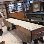 TABLE GAMES: Tracey Beck, co-owner and chief operating officer of The Beck Cos., shows off a shuffleboard table and a pool table, both designed and built by Great American Recreation Equipment Inc., one of six companies under The Beck Cos. umbrella that produces its products at the manufacturer’s 85,000-square-foot facility in North Smithfield. PBN PHOTO/MICHAEL SALERNO