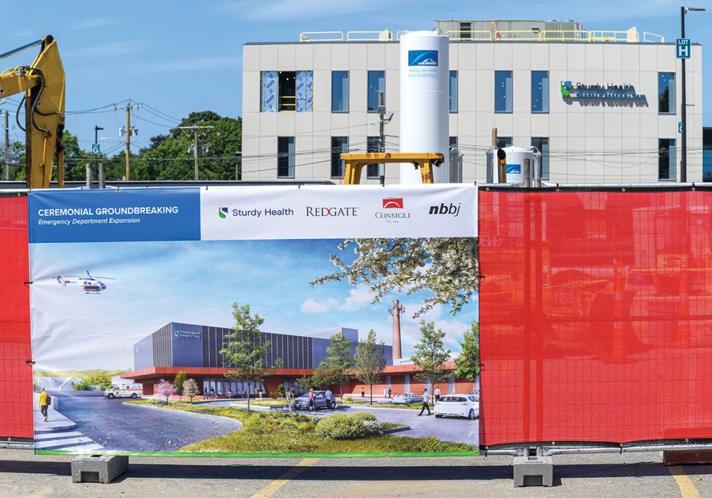 DO IT AGAIN: A recently completed 60,000-square-foot cancer and specialty care building at Sturdy Memorial Hospital in Attleboro peeks over a sign marking the groundbreaking ceremony for a 38,000-square-foot emergency department expansion in August.
PBN PHOTO/MICHAEL SALERNO