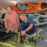 CHUGGING ALONG: Matt Bosgraaf, right, owner of the Kayak Centre in North Kingstown, adjusts the pedal drive system on a fishing kayak in the store’s showroom. With him is John Van Ness, the store’s product specialist. Bosgraaf says the business is growing, helped by the recent acquisition of a kayak rental business.  PBN PHOTO/­ELIZABETH GRAHAM