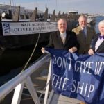 HOPE FLOATS: From left: Thomas Kelly, president and CEO of BankNewport, Tom Goddard, chairman of the Tall Ships Rhode Island Fundraising Committee, and Bart Dunbar, president of TSRI, pose with a “Don’t Give Up the Ship” banner, symbolic of the battle flag flown by Commodore Oliver Hazard Perry during the War of 1812. The hull of the boat is pictured in the background. / 