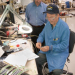 PIECING IT TOGETHER: Mission equipment assembler Joe Ramos, seated, pieces together a component to a Raytheon weapons system while process engineer William Cicchelli looks on. / 