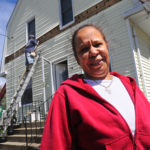 KEEPING IT IN HOUSE: Cranston resident Rita Bisono stands in front of her home while a Rhode Island Insulation worker starts the process of insulating it. / 