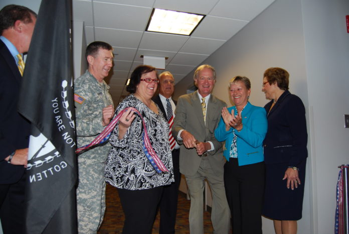 At today's dedication of the new T.F. Green Airport military lounge, R.I. Airport Corp. President & CEO Kelly Fredericks is hiding behind the flag, next to Adjudent Gen. Kevin McBride, Warwick Councilwoman Camille Vella-Wilkinson, Warwick Councilman Edgar Ladouceur, Gov. Lincoln Chafee, R.I. Military Organization Vice Chair Lori Ashness and RIAC Board Chair Dr. Kathleen Hittner. / COURTESY R.I. AIRPORT CORPORATION