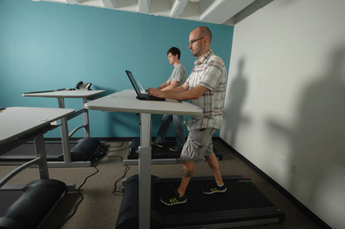 WALKING THE TALK: ShapeUp employees Jake Denham, left, and Andrew Bianchi get in exercise while doing their jobs at stand-up desks in the company’s Providence offices. / PBN PHOTO/BRIAN MCDONALD