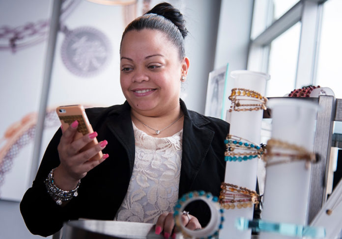 CROWN JEWEL: Alex & Ani employee Keisy Marcano checks messages on her smart-phone at the retail store in Cranston. The company has adopted new sensor technology called the iBeacon that allows stores to detect, send messages and interact with customer mobile phones when they are in or near the store. / PBN PHOTO/MICHAEL SALERNO