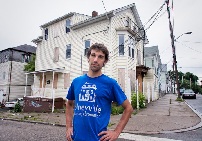 CAN’T GO HOME: Chris Ackley, stewardship manager at Olneyville Housing Corp., in front of 238 Amherst St. in Olneyville. The three-family home has yet to be foreclosed on because banks don’t want to own it. / PBN PHOTO/MICHAEL SALERNO
