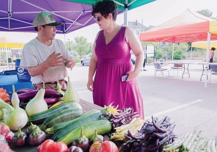 FIRM ROOTS: Blia Moua speaks with Rachel Newman Greene, director of partnerships and community projects for West Elmwood Housing Development Corporation, at a farmers market in the organization’s parking lot. / PBN PHOTO/MICHAEL SALERNO