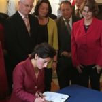 GOV. GINA M. RAIMONDO signs the executive order establishing a group that will restructure Medicaid in Rhode Island. She is surrounded by members of the group. Elizabeth H. Roberts, state secretary of health and human services, is standing behind Raimondo. / PBN PHOTO/ELI SHERMAN