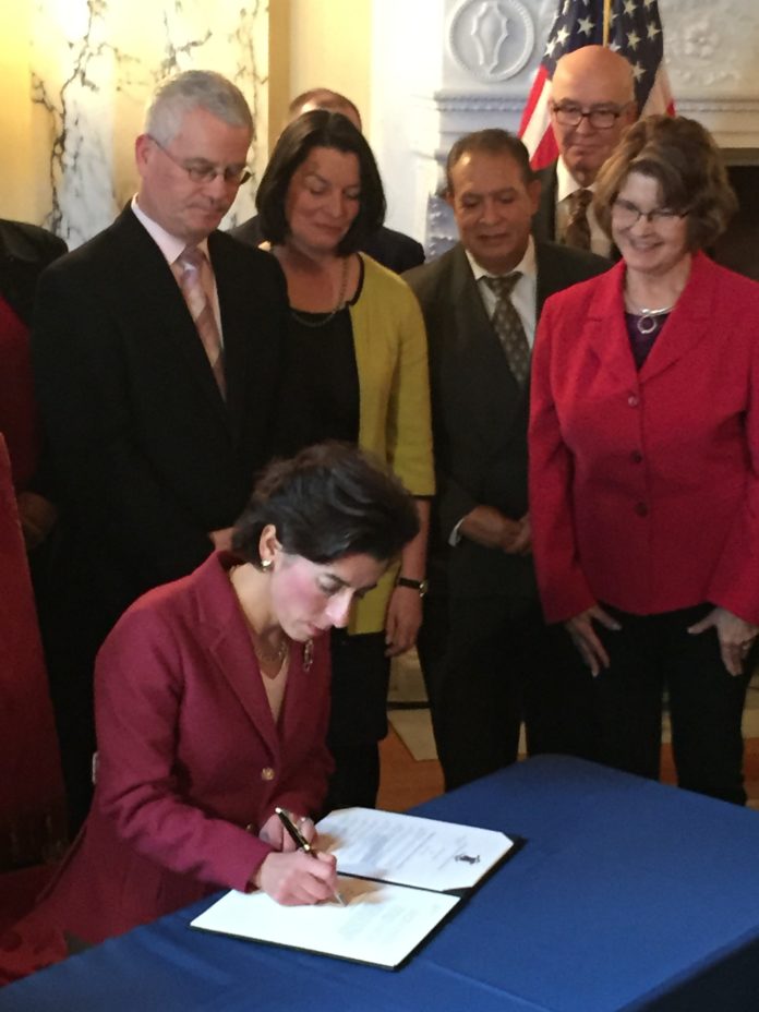 GOV. GINA M. RAIMONDO signs the executive order establishing a group that will restructure Medicaid in Rhode Island. She is surrounded by members of the group. Elizabeth H. Roberts, state secretary of health and human services, is standing behind Raimondo. / PBN PHOTO/ELI SHERMAN