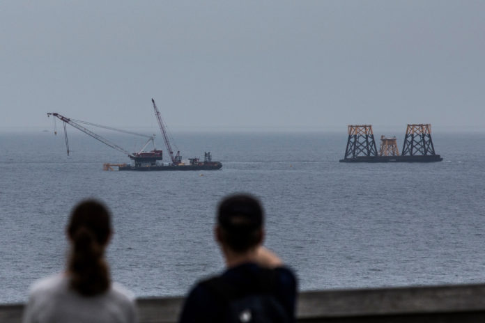 PEOPLE LOOK out at a Deepwater Wind LLC offshore wind farm under construction off the coast of Block Island on Monday. Deepwater Wind LLC is installing a massive steel frame, more than 1,500 tons, that sits on the seabed and juts about 70 feet from the water south of Rhode Island. By the end of next year there will be five of these platforms, each supporting a huge turbine, the first offshore wind farm in U.S. waters. / BLOOMBER NEWS/SHIHO FUKADA