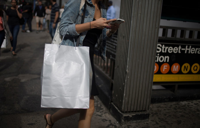 COMMERCE DEPARTMENT DATA released Monday showed that household spending rose in August more than expected, at the same time that July levels were revised higher than preciously reported. A woman checks a mobile device while carrying a shopping bag on 34th Street in New York on Saturday. / BLOOMBERG NEWS PHOTO/JOHN TAGGART
