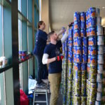 Kristine Christensen, left, and Pierson Booher, both employees of Durkee, Brown, Viveiros & Werenfels Architects of Providence, build the indoor seawall for the sculpture &ldquo;Turning the Tides On Hunger&rdquo; with their teammates. The event March 12 involved six teams and took place at the Providence Place Skybridge Concourse during Canstruction Build Day to collect canned food for the Rhode Island Community Food Bank. / COURTESY RHODE ISLAND COMMUNITY FOOD BANK