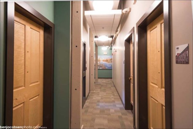 NEW USE: The original support posts for the factory floor are visible at left and now support the apartment corridor walls. Individual apartments are accessed off the hallway. At right, an image of the mill is used on a room number plate. At the end of the hallway, an original painting donated by artist Anthony Tomaselli is visible.
