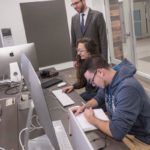 MEDIA HUB: Adam David Roth, standing, interim director of the Harrington School of Communications at URI, speaks with Amber Beech, of Warwick, a junior majoring in communication studies, in the university's new media hub. Nathan Finnigan, of Coventry, a senior majoring in psychology, works at right. / PBN PHOTO/MICHAEL SALERNO