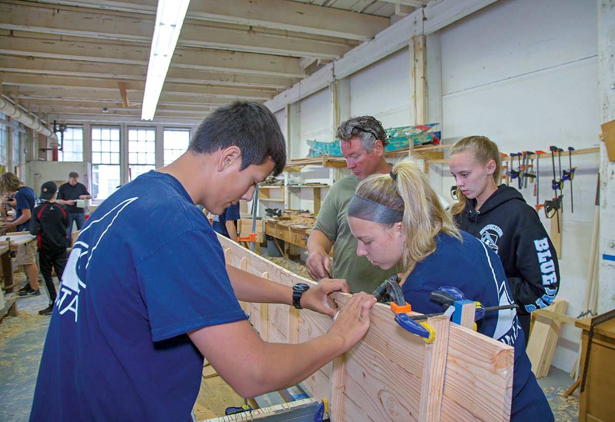 SKIFF WORK: Kirk Cusic, green shirt, executive director of the Herreshoff Marine Museum in Bristol, works with students on a traditional Westport skiff, as part of the Rhode Island Marine Trades Association’s Youth Summer Boatbuilding Program.  / COURTESY RIMTA/BILLY BLACK