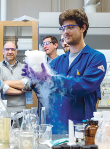 LAB WORK: From left, Brenton DeBoef, URI professor of chemistry, and Alcinous Pharmaceutical team members David Worthen, chief operations officer, and Benjamin Barlock, chief technology officer, observe URI graduate student David Robinson in DeBoef’s lab in the Beaupre Center at URI. / PBN PHOTO/RUPERT WHITELEY