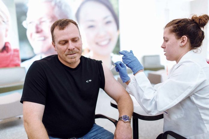 COMPANY PRIORITY: Jeff Guilbert, product-quality leader at Amgen Rhode Island in West Greenwich, receives a flu shot from University of Rhode Island pharmacy student Kate Syder during one of the company’s in-office clinics this past fall. / PBN PHOTO/RUPERT WHITELEY COMPANY PRIORITY: Jeff Guilbert, product-quality leader at Amgen Rhode Island in West Greenwich, receives a flu shot from University of Rhode Island pharmacy student Kate Syder during one of the company’s in-office clinics this past fall. / PBN PHOTO/RUPERT WHITELEY