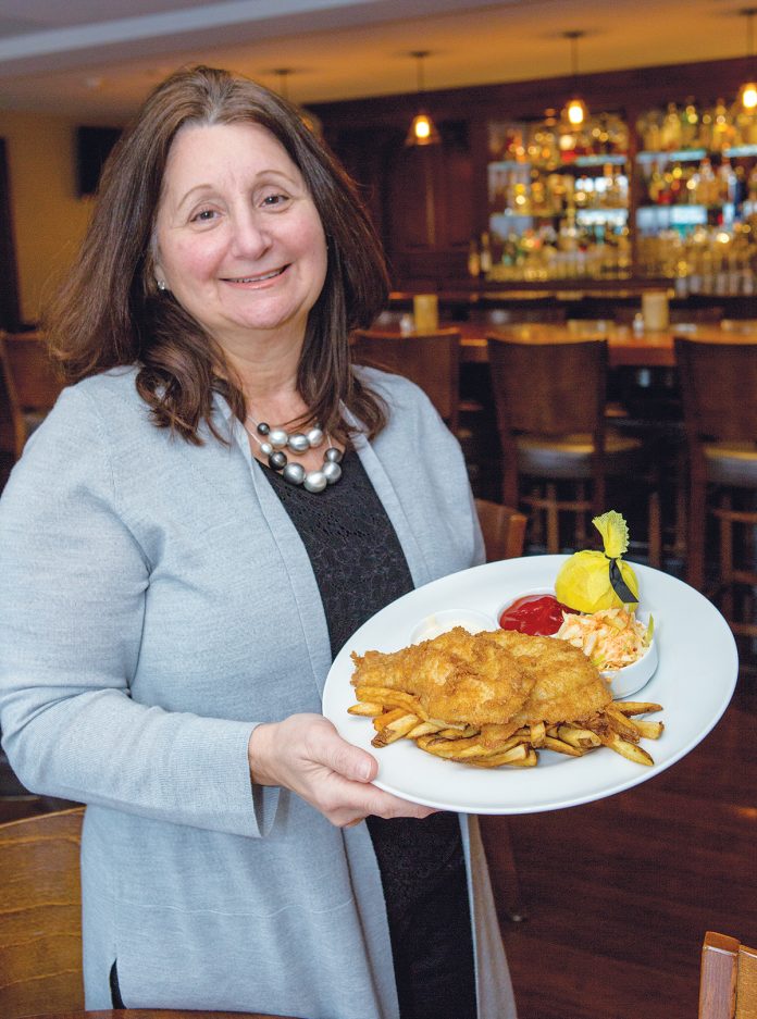 AGE-OLD FAVORITE: Joann Carlson, owner of Jo’s American Bistro in ­Newport, hold’s a plate of the restaurant’s famous fish and chips, a 50-year-old family recipe. / PBN PHOTO/KATE WHITNEY LUCEY
