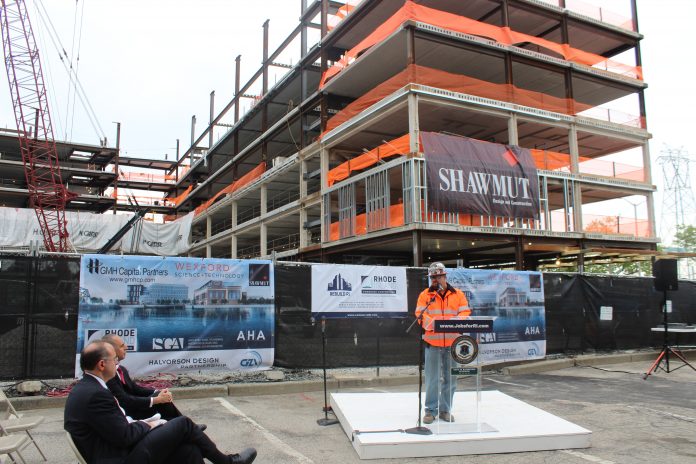 JAMES WOODWARD, superintendent at Shawmut Design and Construction, speaks during the topping off ceremony for the River House apartments project, in which the final steel beam is placed atop the structure. Seated are Commerce Secretary Stefan Pryor, foreground, and Providence Mayor Jorge O. Elorza. / COURTESY SHAWMUT