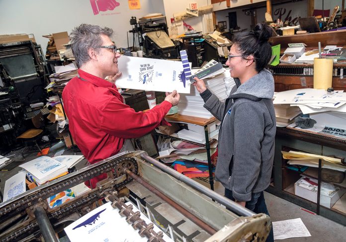 LASTING LETTERPRESS: Dan Wood, owner and founder of the Dan Wood Rhode Island letterpress print shop, works with Lois Harada, shop manager, on the Polyman 50 automatic-fed cylinder letterpress at the shop’s headquarters in Providence. / PBN PHOTO/MICHAEL SALERNO