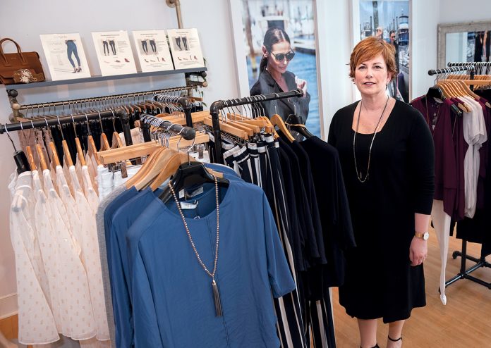 POPULAR DESIGNS: Robin Barrett Wilson, CEO and founder of robin b., a 3-year-old women’s fashion design and boutique in Cranston, wears one of her designs, a black sleeveless sheath dress underneath a cardigan sweater, both made in New York. The outfit on the rack next to her has a Lola & Sophie top and Parkes Smith jeans.  / PBN PHOTO/MICHAEL SALERNO