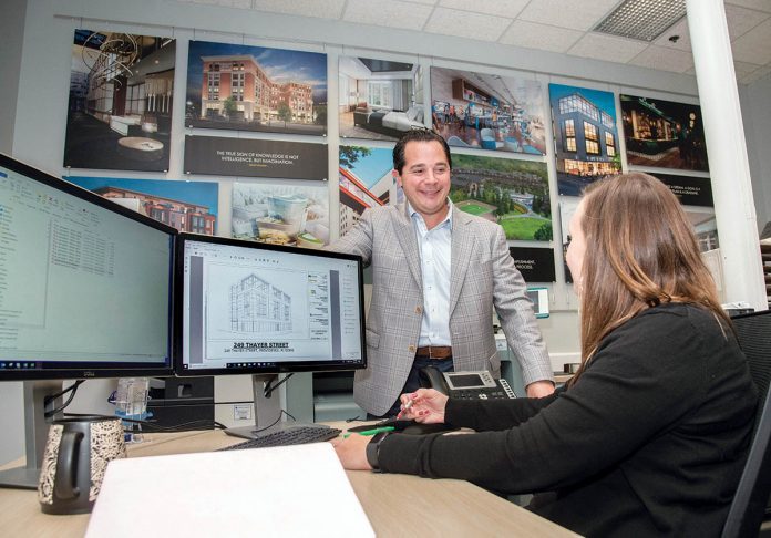DEDICATED DESIGN: Eric Zuena, managing principal at ZDS, speaks with Julie Jancewicz Bartlett, project manager/associate. The wall behind Zuena features designs from many of the company’s clients.  / PBN PHOTO/MIKE SALERNO