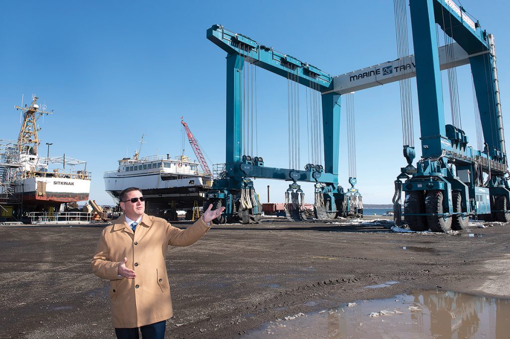 TRAVEL LIFT: Quonset Development Corp. Managing Director Steven J. King talks about the J. Goodison Co. Shipyard on the waterfront at the Quonset Business Park in North Kingstown. The company boasts having the largest travel lift in New England, which can be seen in the background.  / PBN PHOTO/DAVE HANSEN