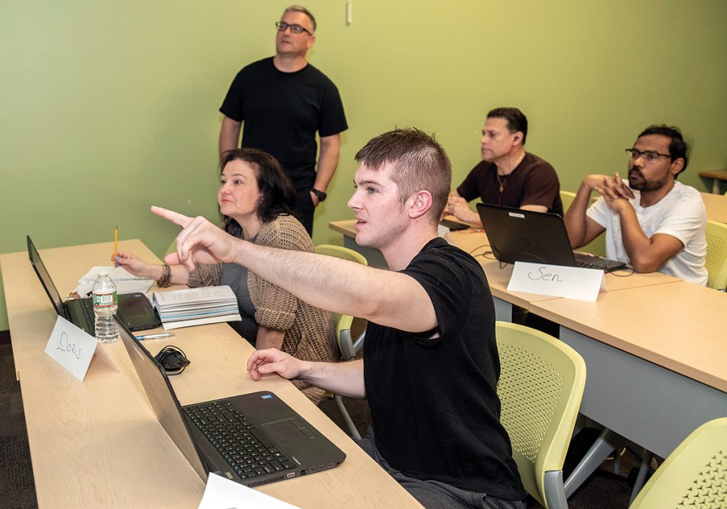 COMPUTER SKILLS: Students participate in a computer skills class at the Cranston Public Library. Chris Lamothe, standing, is teaching the classes in Cranston while the Providence Public Library is being renovated, then he will teach at PPL. Students, from left, are Doris Jaramillo of Pawtucket; Tom Lanigan, foreground, of East Providence; Arnoldo Casillas of Providence; and Senthil Muthiya of Cranston, all of whom are employed.  / PBN PHOTO/MICHAEL SALERNO