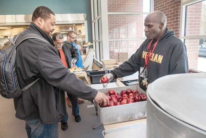 BUSY KITCHEN: Kevin Burton, right, a staffer in the Amos House soup kitchen in Providence, offers an apple to Kashaka Williams of Providence. Amos House President and CEO Eileen Hayes said the soup kitchen is the largest in the state and serves between 120,000 and 130,000 meals annually.  / PBN PHOTO/MICHAEL SALERNO