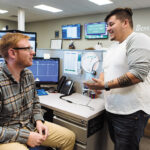 SHOP TALK: Vertikal6 service desk apprentices Steven Walsh, left, and Dylan Herrera share a conversation in the company’s office. / PBN PHOTO/RUPERT WHITELEY